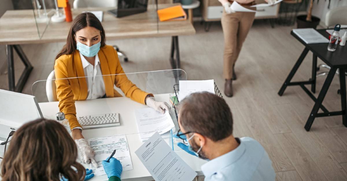 woman sitting at a desk across from a man and lady. all three are wearing surgical mask and gloves